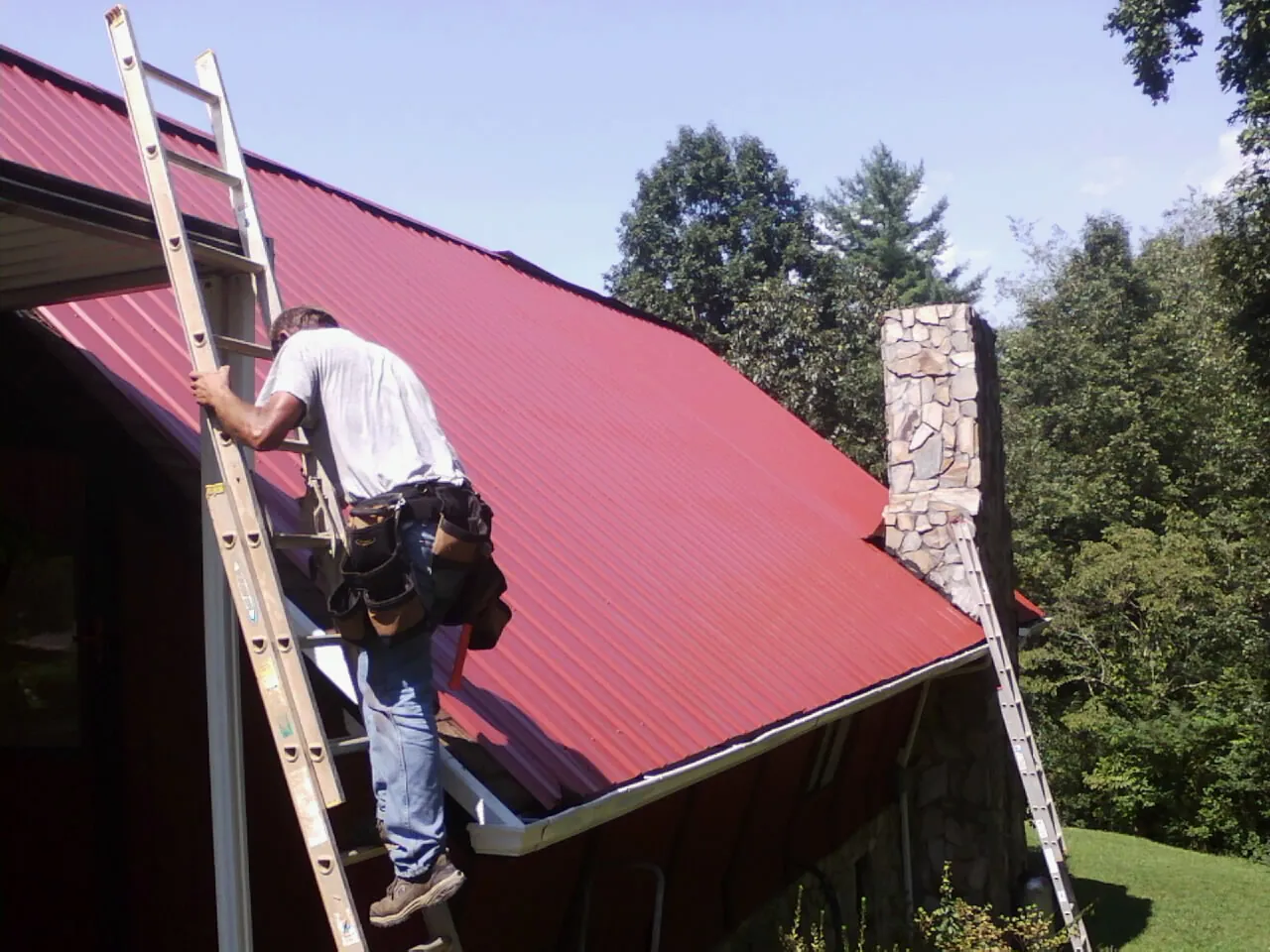 Roofer on a ladder beside a red metal roof and stone chimney — Soffit & Fascia Repair by Roofing Brothers in Deal Island